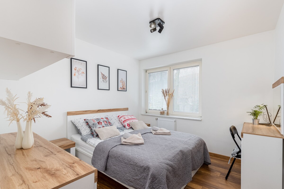 A calming bedroom features a spacious bed dressed in a gray quilt and decorative pillows. Framed artwork adorns the walls, while a window allows natural light to illuminate the area. A small table and chair are positioned nearby, creating a functional space.