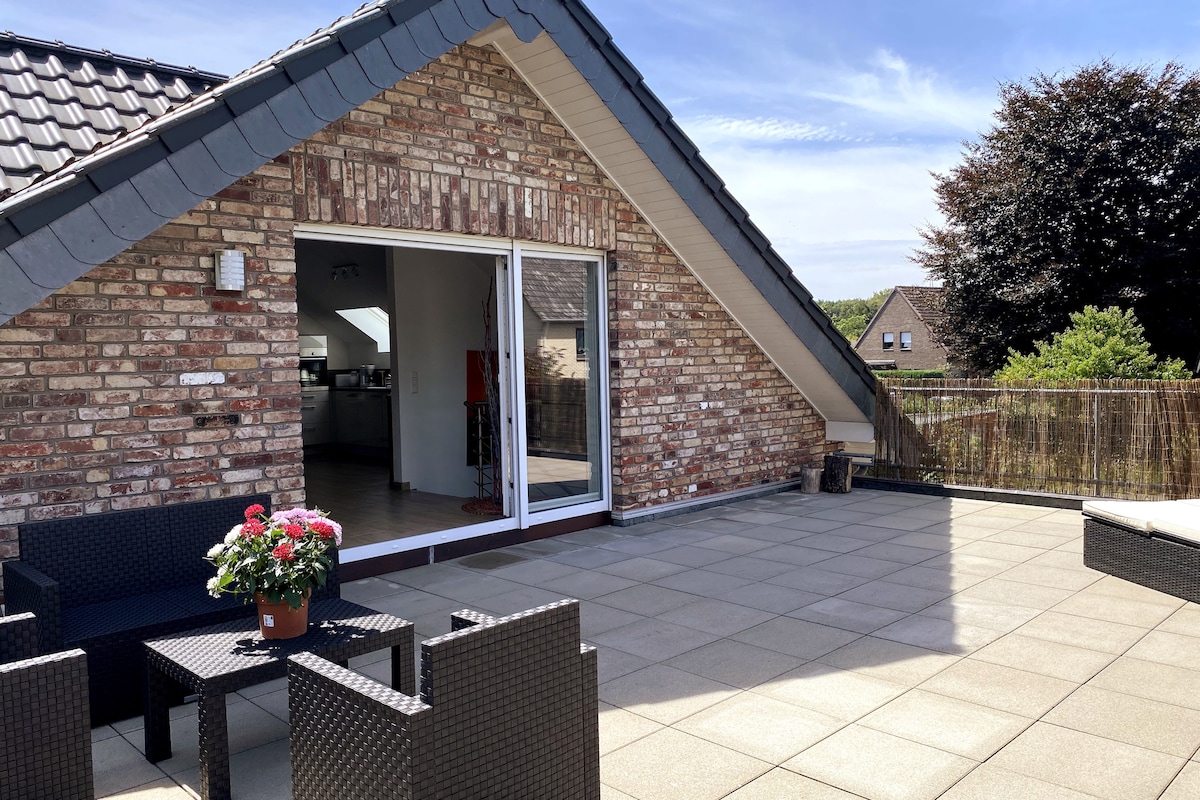 A spacious terrace features a tiled surface and is accented by a potted flower arrangement. Comfortable seating is arranged around a small table, while large glass doors lead into the interior, providing a seamless connection to the outdoor space.