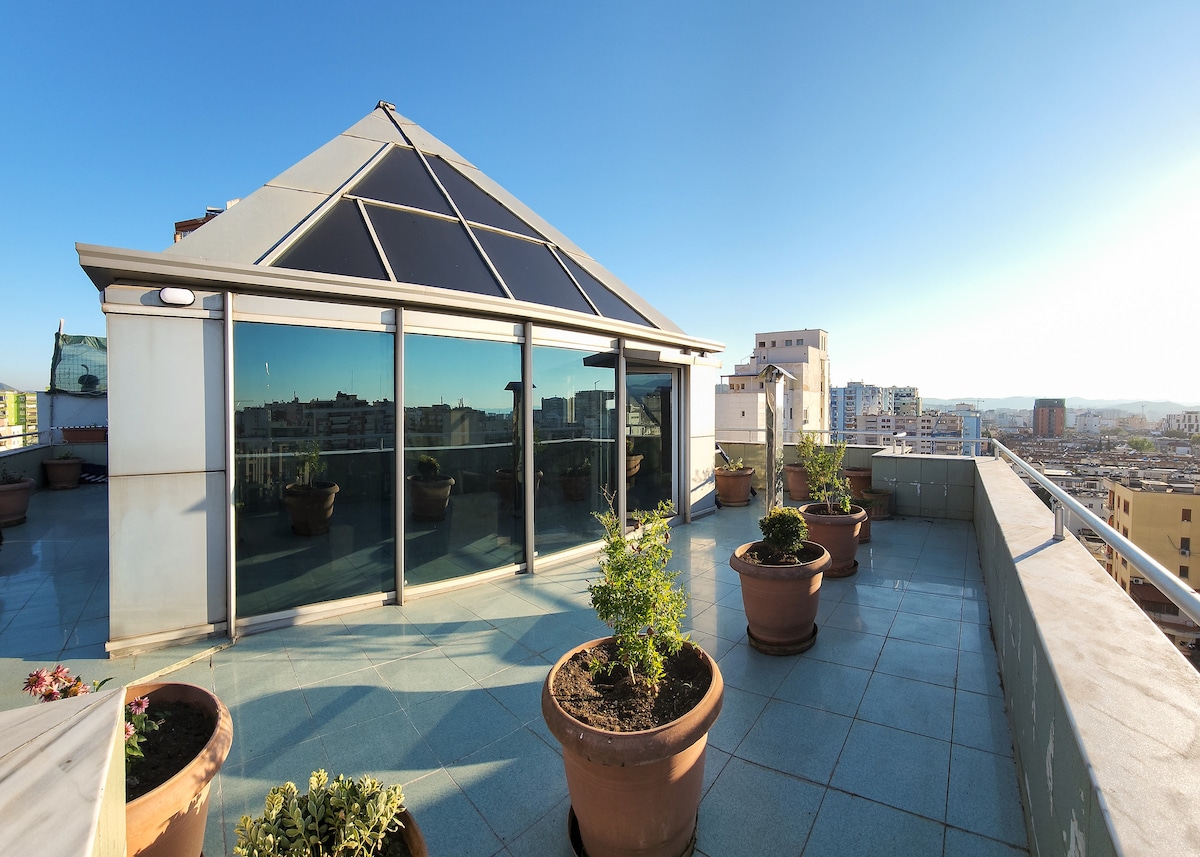 A spacious balcony surrounding the glass pyramid features several potted plants, creating a serene outdoor space. The large glass walls reflect the cityscape, offering panoramic views, while a clear blue sky enhances the inviting atmosphere of the terrace.