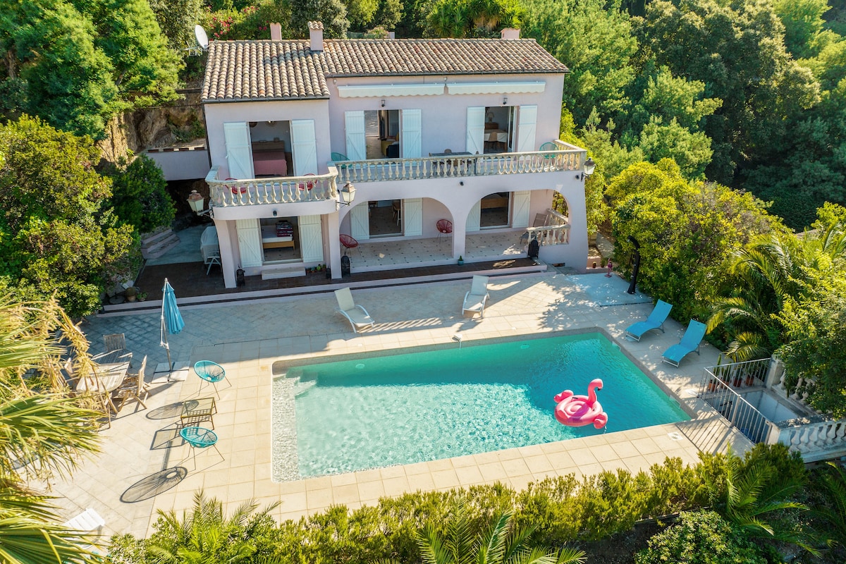 An aerial view of a charming villa surrounded by lush greenery. A private pool with a floating flamingo is featured in the foreground, complemented by sun loungers and outdoor seating. The spacious terrace and balcony offer additional outdoor living areas, enhancing the villa's inviting appearance.