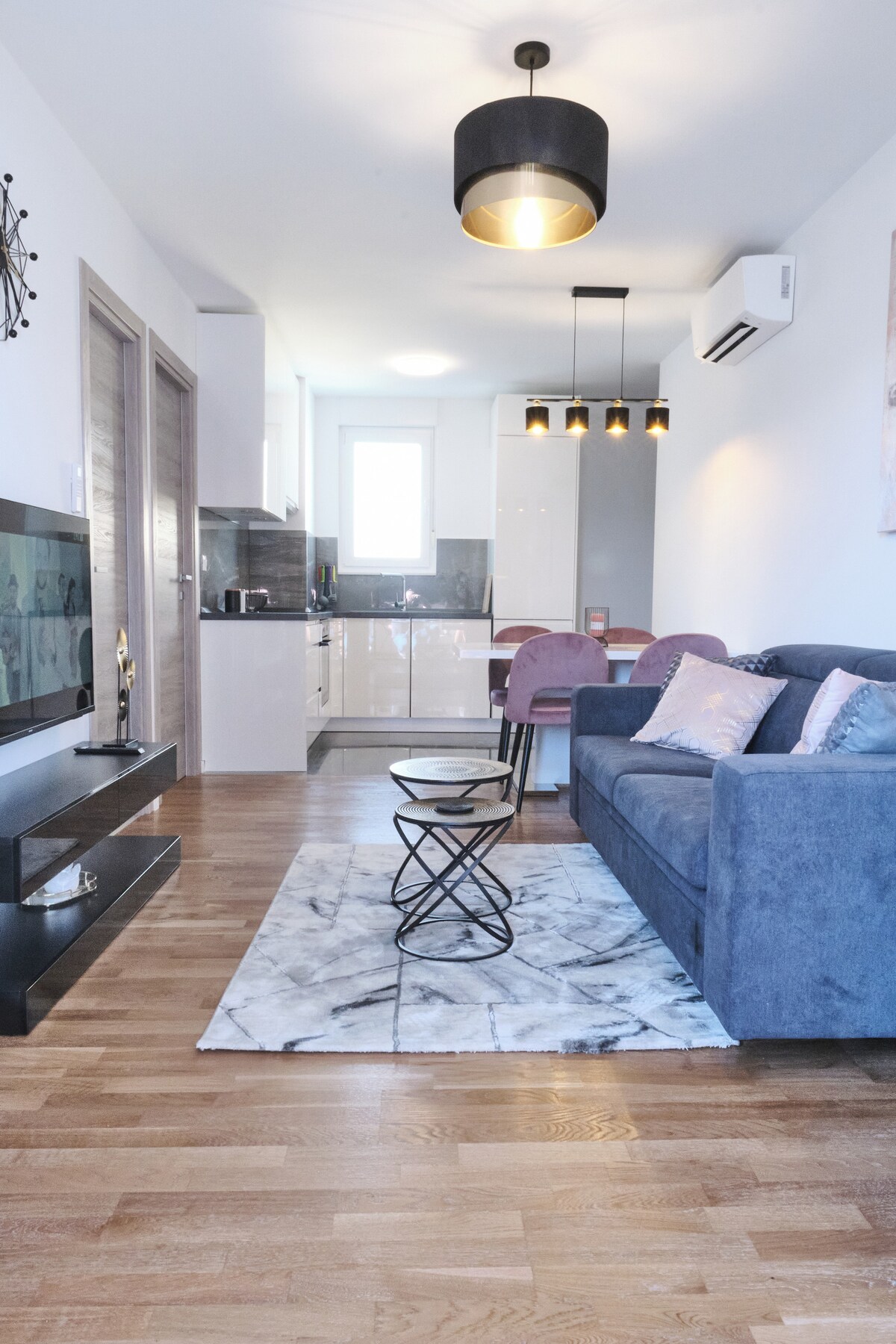 A modern living area features a comfortable blue sofa complemented by decorative pillows. A stylish area rug lies beneath a small black table. An entertainment unit is positioned across from the sofa, and a well-equipped kitchen area is visible in the background, illuminated by pendant lighting.