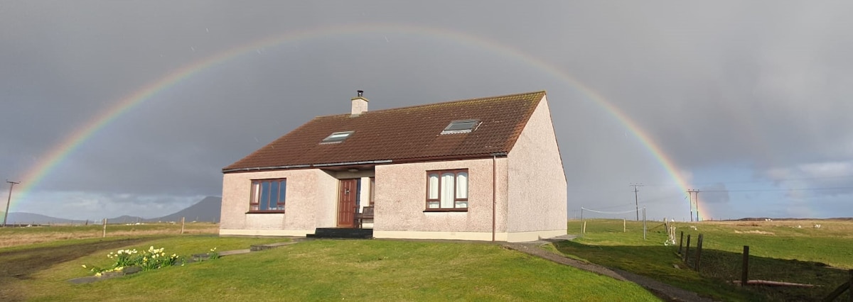 A single-story cottage is set against a backdrop of open fields, featuring a sloped roof and evenly spaced windows. A vibrant rainbow arches over the home, while a portion of the garden with blooming flowers is visible in the foreground.