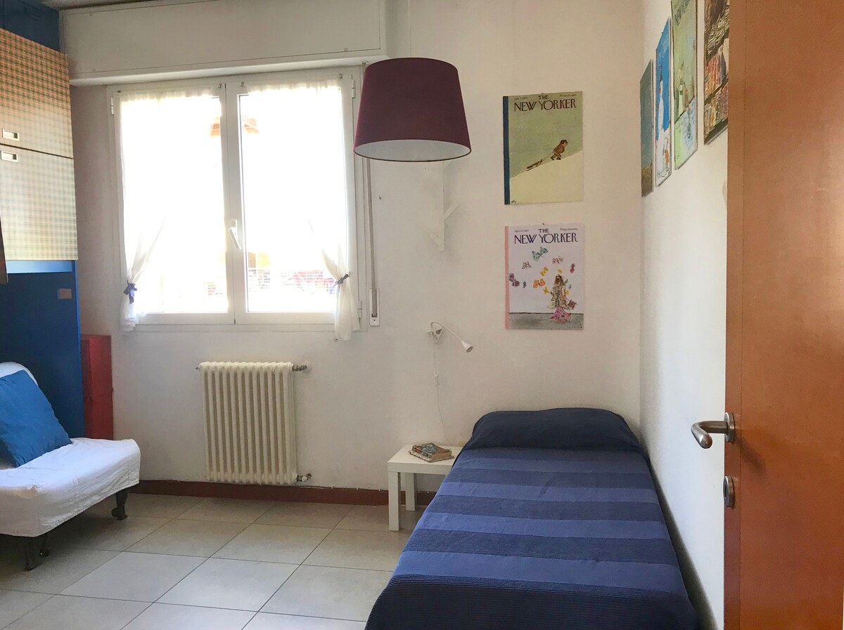 A simple room features a single bed dressed in a blue striped cover. A chair is positioned beside the bed, while a small white side table holds a lamp. Natural light enters through two windows covered with light curtains, and artwork is displayed on the wall.