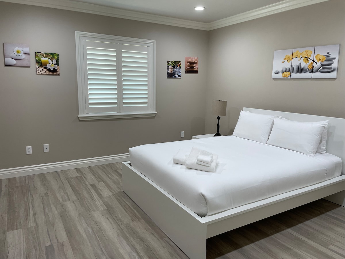 A well-lit bedroom features a spacious bed with crisp white linens and towels neatly arranged at the foot. Light-colored walls are adorned with three tranquil wall art pieces, complemented by a bedside lamp and a window with decorative shutters.