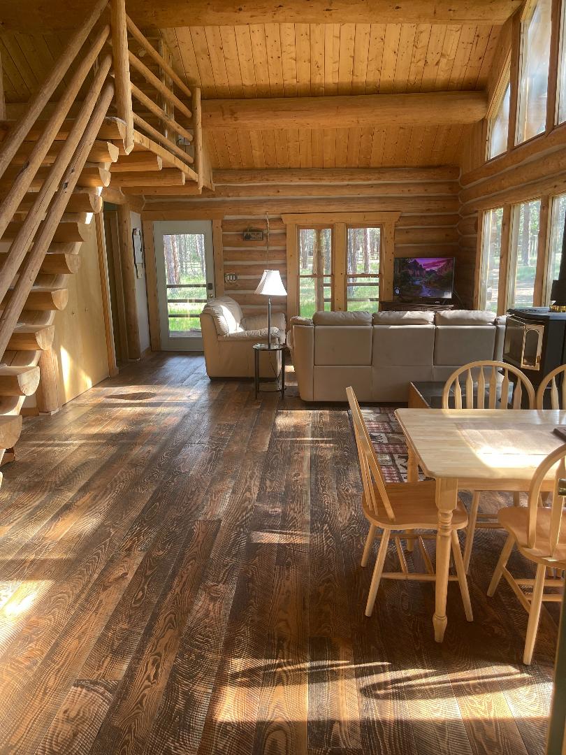 An open living area features a wooden staircase leading to the upper level. A cozy seating arrangement and a television are positioned near large windows, allowing natural light to fill the space. A dining table with four chairs is situated adjacent to the seating area.