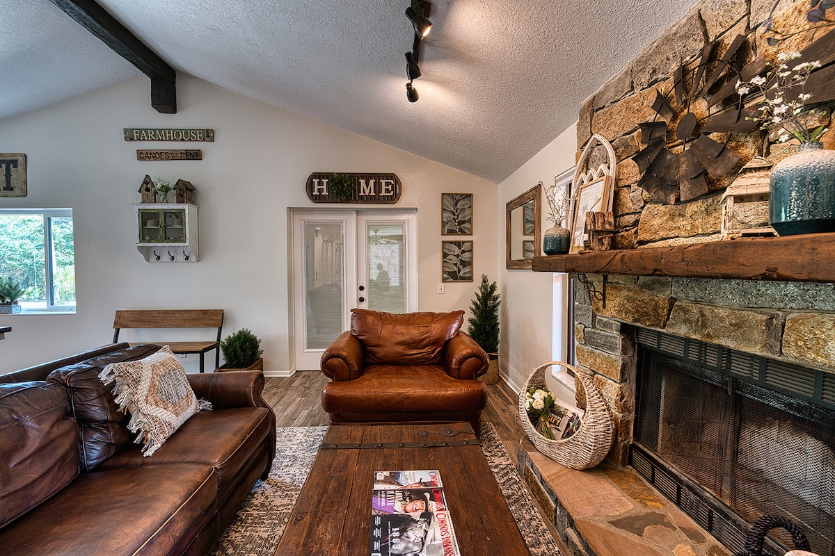 A welcoming living area is featured with leather sofas and a wooden coffee table. A stone fireplace is adorned with decorative elements, while natural light enters from nearby windows. The rustic decor includes wall art and plants, enhancing the cozy environment.
