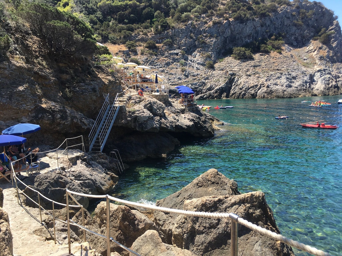 A rocky shoreline meets clear turquoise waters, with steps leading to a small beach area. Sun umbrellas provide shade for visitors enjoying the sun. Nearby, kayakers navigate the serene waters, while lush greenery and rugged cliffs frame the picturesque scene.