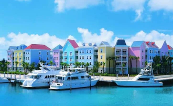 A vibrant waterfront scene features colorful buildings in pastel hues of blue, pink, and yellow. Moored yachts are visible in the serene turquoise waters, with a clear blue sky and soft clouds creating a bright backdrop.