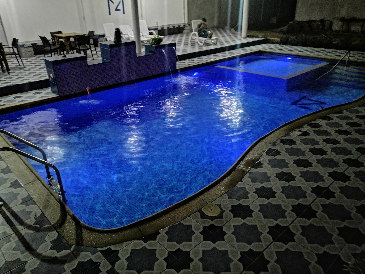 A well-lit swimming pool is surrounded by patterned tiles in various shades. The water is a vibrant blue, reflecting soft lighting from above. A seating area and lounge chairs are visible in the background, offering spaces for relaxation.