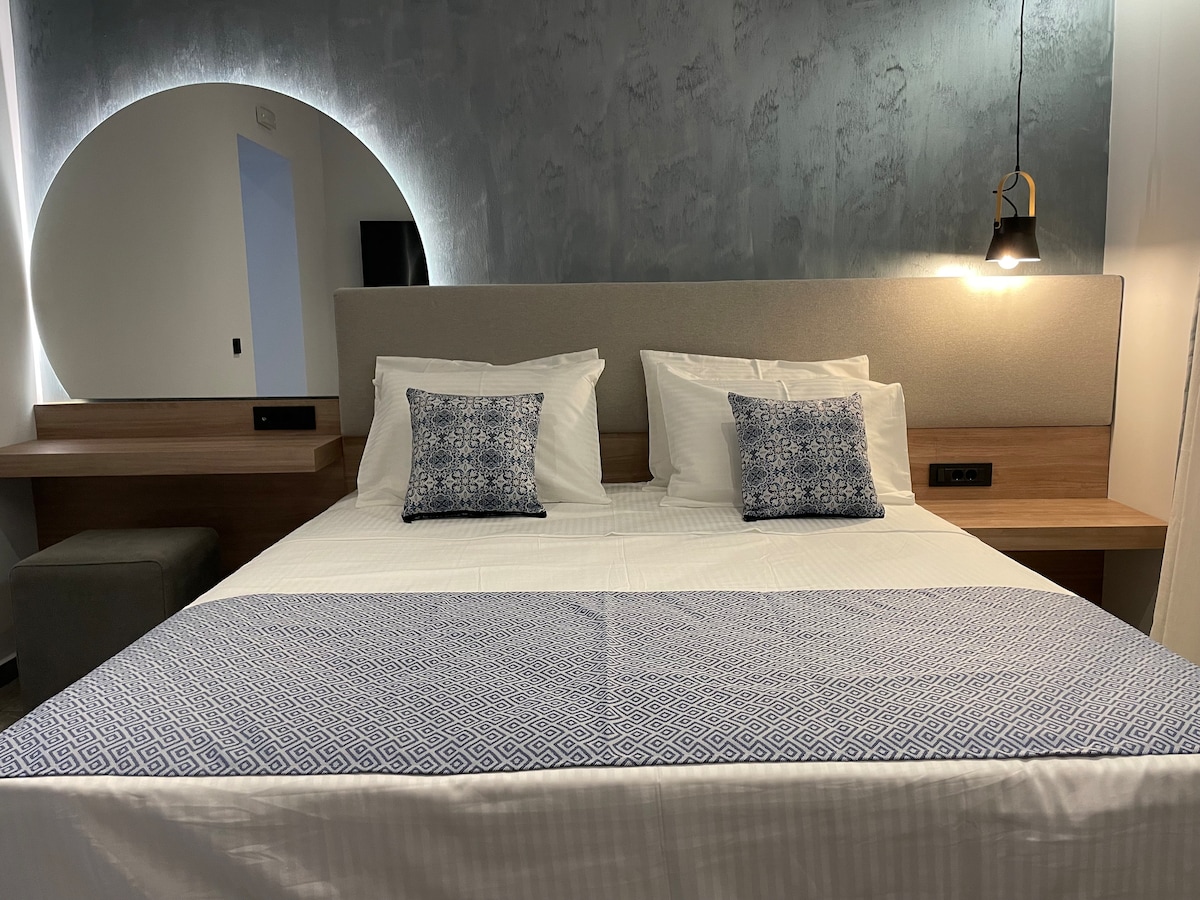 A comfortable sleeping area is highlighted by a spacious bed adorned with decorative pillows. The minimalist design includes a stylish headboard and a wooden accent wall, complemented by a soft ambient light fixture. A small seating area is visible beside the bed.