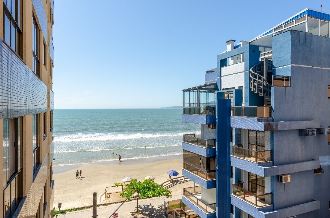 A coastal view is presented from the balcony, showcasing the ocean waves and sandy beach below. The image captures neighboring buildings, with balconies and beach umbrellas visible along the shore where individuals can be seen enjoying the seaside.