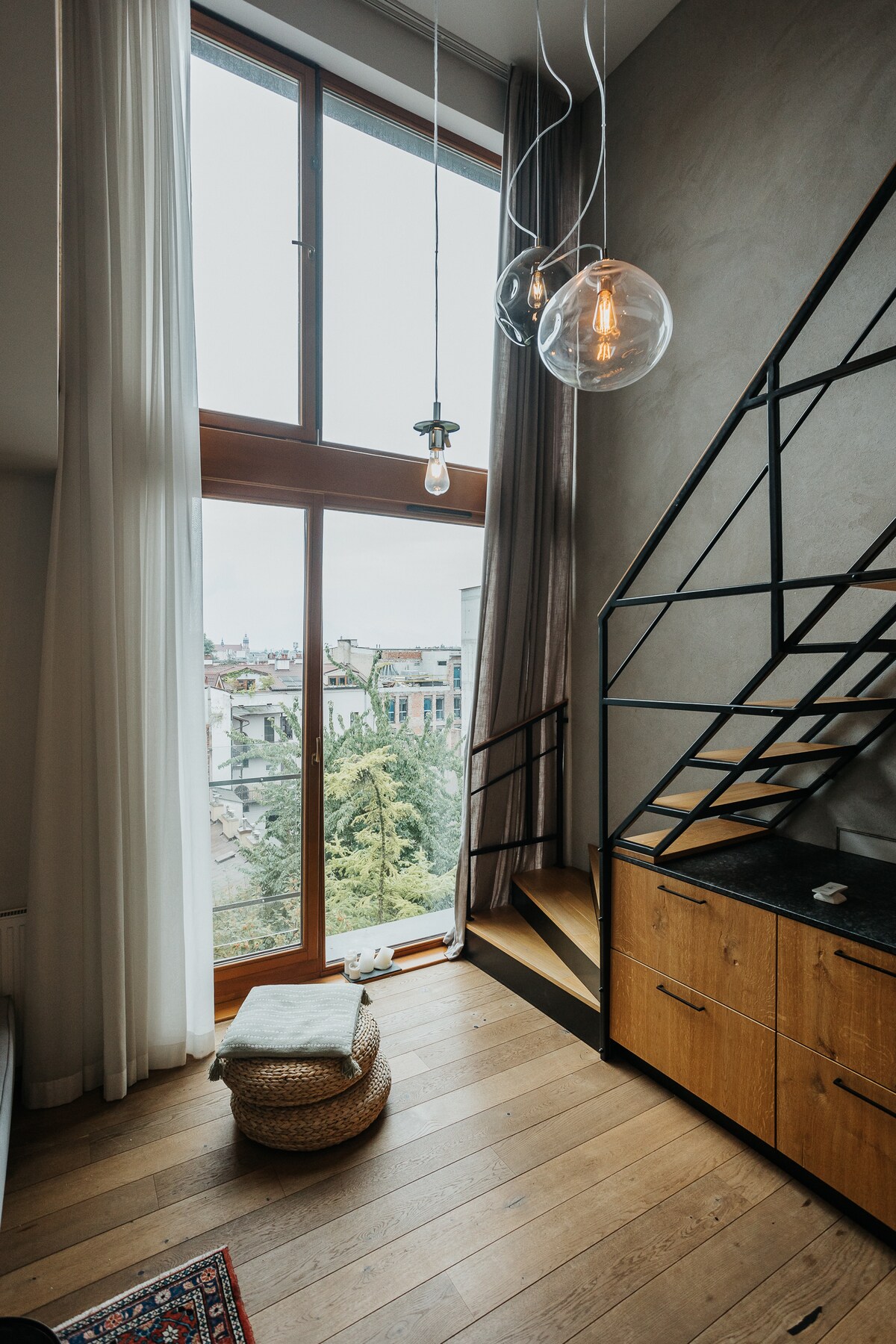 A corner of the apartment features large windows that allow natural light to fill the space, revealing a view of the city. An elegant staircase leads to the upper level, accompanied by decorative hanging lights. A woven pouf sits on the hardwood floor.