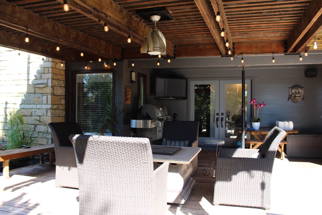 A private deck is surrounded by wooden beams and features overhead string lights. Comfortable seating is provided by four wicker chairs and a table in the center. A BBQ is positioned on one side, with large glass doors leading indoors.