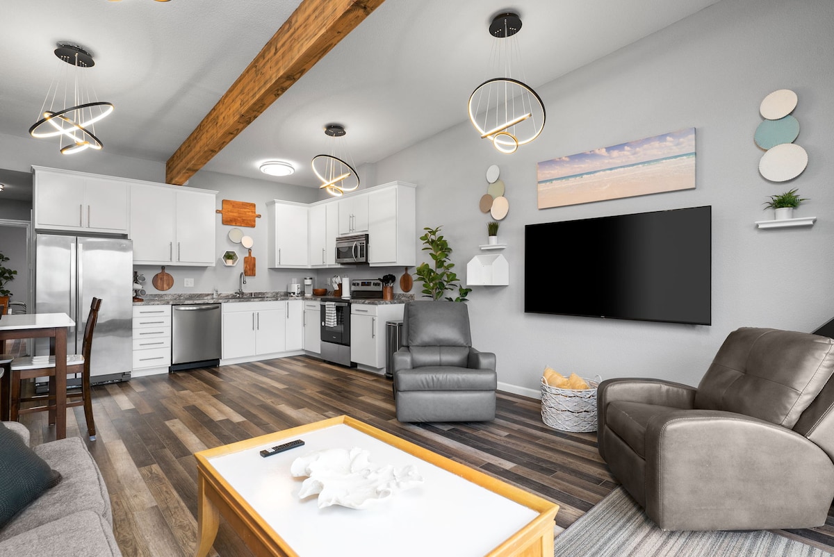 An open concept living space is shown, featuring light-colored cabinets, stainless steel appliances, and a large television mounted on the wall. A dining area is visible, along with comfortable seating arrangements, including a gray sofa and two armchairs. Unique lighting fixtures hang above, complementing the wood beam highlights.
