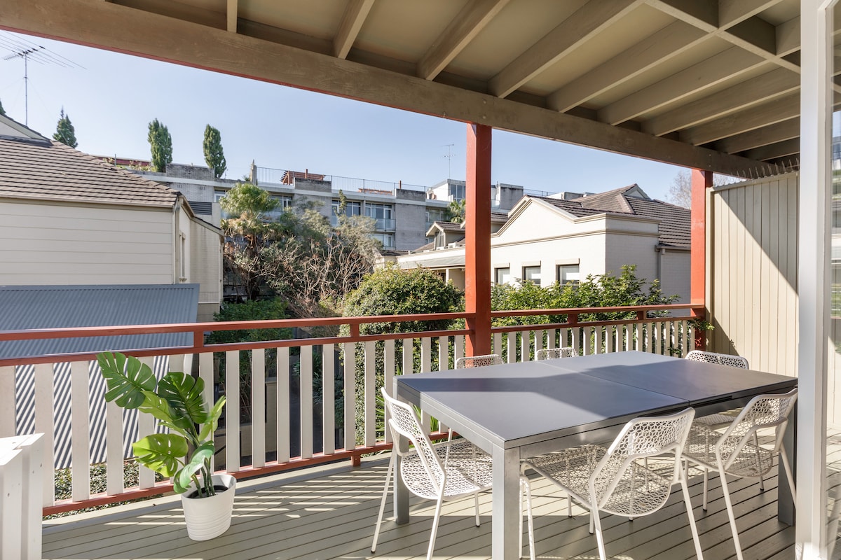 A covered outdoor balcony offers a dining table with a neutral tabletop and white chairs. Lush greenery is visible in the surrounding area, with sunlight illuminating the space. Nearby buildings provide a glimpse of the neighborhood, enhancing the private outdoor atmosphere.