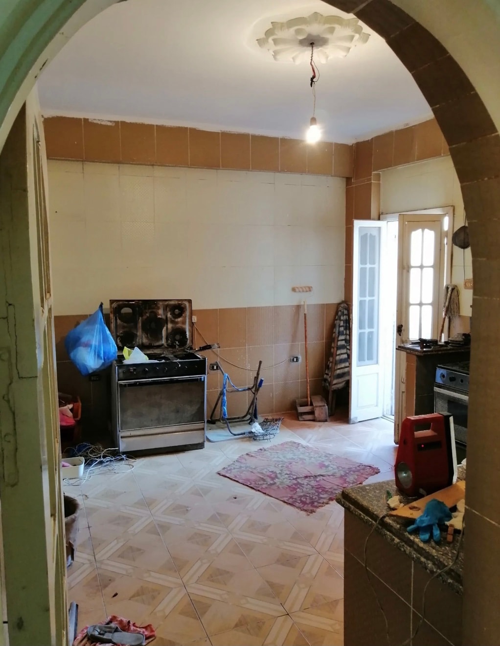 A kitchen is shown with a simple layout, featuring a stove positioned against a tiled wall. Natural light floods in from a partially open door, highlighting a patterned rug on the floor. Various tools and cleaning items are placed on the countertop.