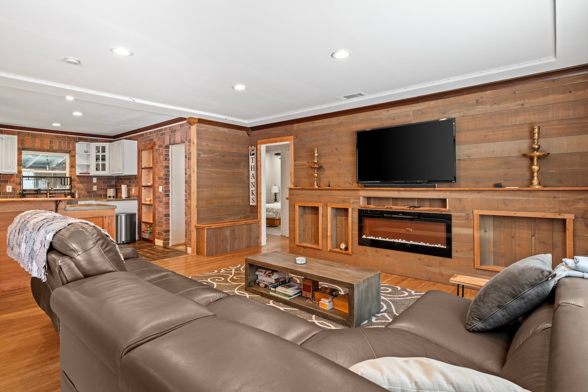 A spacious living area features warm wooden accents and a modern sectional sofa. A flat-screen TV is mounted above a contemporary fireplace. The full kitchen is visible in the background, providing easy access for cooking and entertaining.