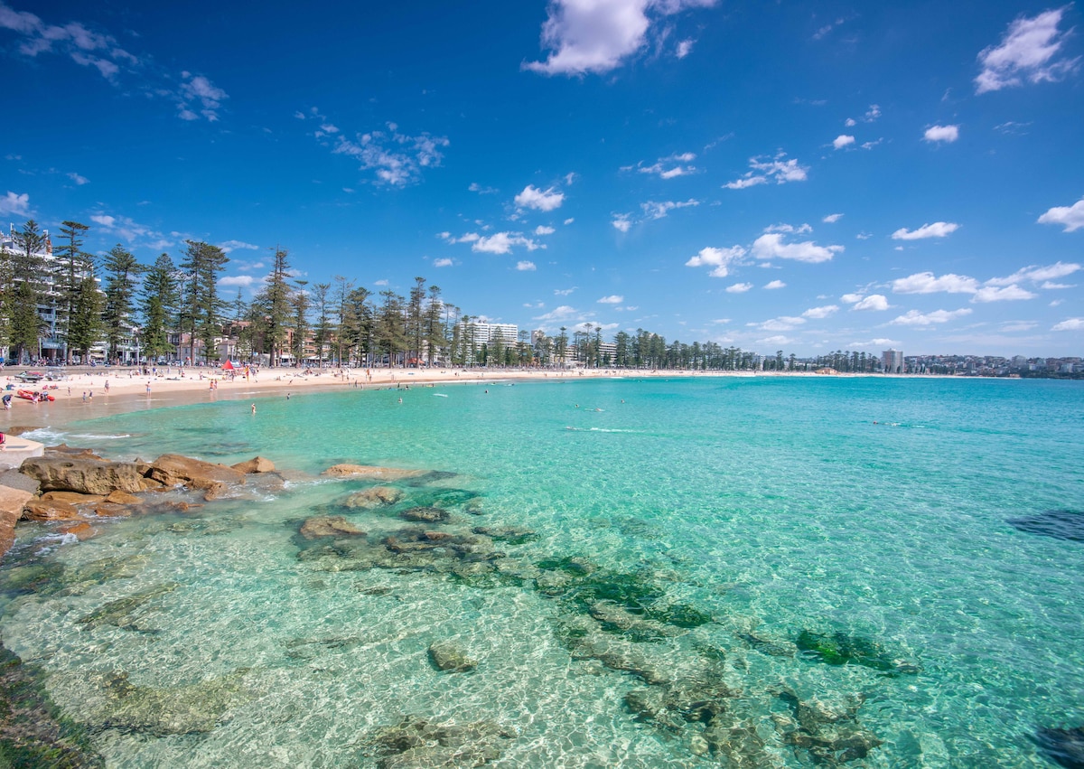 A serene view of a clear, turquoise ocean extends towards a sandy beach lined with people enjoying the sun. Rocky formations are visible in the water, while palm trees and buildings form a coastline backdrop under a bright blue sky with scattered clouds.