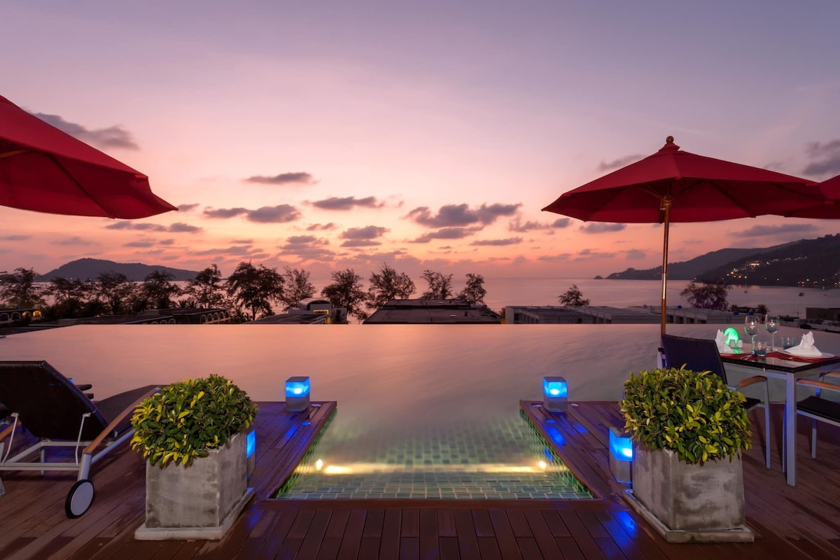 An infinity pool extends seamlessly towards the horizon, offering a panoramic view of the sunset. Comfortable lounge chairs are positioned nearby, shaded by large red umbrellas. Soft lighting accents the pool's edge, creating a serene atmosphere against the backdrop of gentle waves and distant hills.