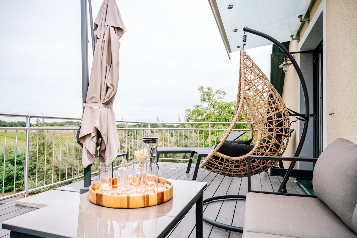 A private terrace is furnished with a wicker hanging chair and a modern table. A beige umbrella provides shade, while a wooden tray holds glassware, suggesting a space for relaxation. Lush greenery and vineyards are visible in the background, enhancing the outdoor setting.