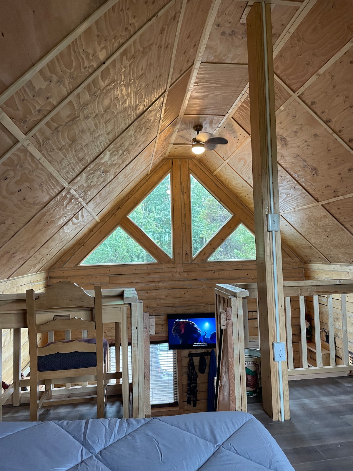 A lofted interior space is shown, featuring a sloped wooden ceiling to enhance an open feel. A triangle-shaped window allows natural light to enter, illuminating a small seating area with two chairs. A television is positioned in front of the seating area.