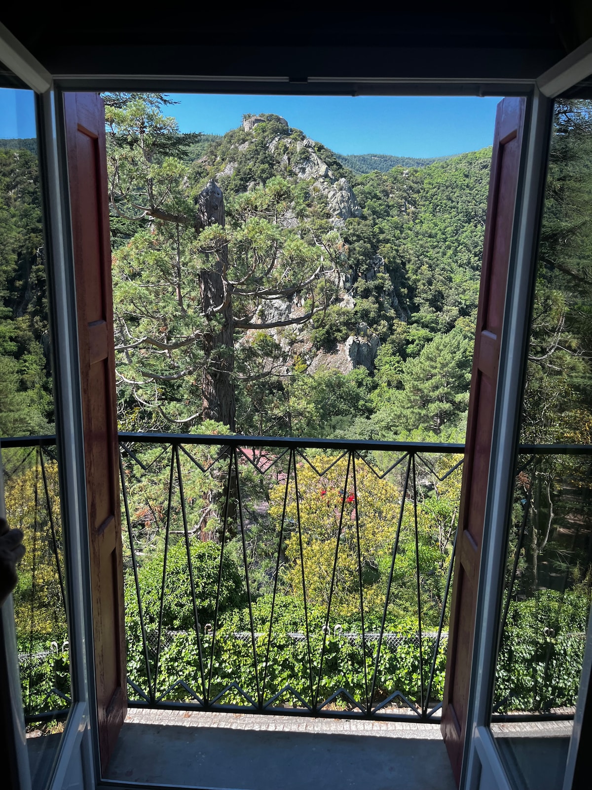 A balcony view showcases dense greenery and a prominent rocky outcrop against a mountainous backdrop. Large windows open to allow natural light, framed by wooden shutters that emphasize the outdoor scenery.