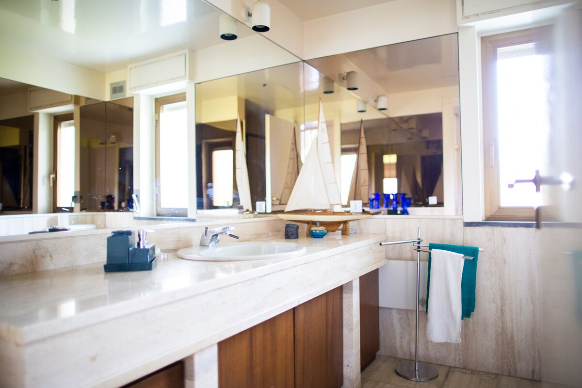 The bathroom features a marble countertop with a spacious sink area. A large mirror reflects natural light from the window, enhancing the room's openness. A decorative model sailboat is displayed on a shelf, accompanied by various toiletries. A towel hangs neatly on a chrome rack.