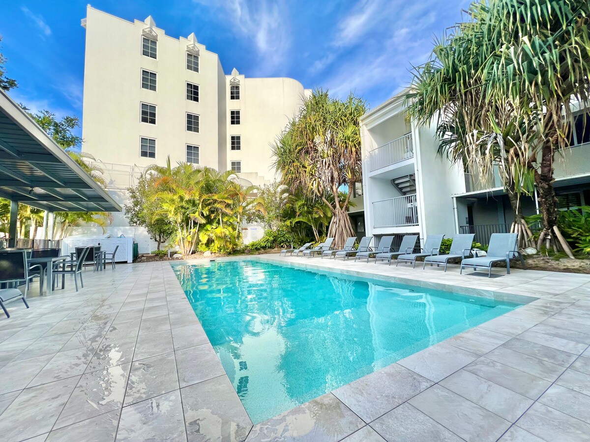 A clear, inviting swimming pool reflects the sky, surrounded by a sun terrace featuring multiple lounge chairs. Lush greenery and palm trees enhance the tropical ambiance, while a multi-story building stands in the background, contributing to the serene environment.