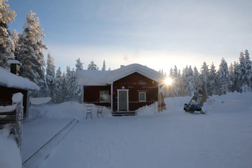 A cozy red cabin is surrounded by snow-covered trees, reflecting a winter scene. Sunlight shines through the forest, casting a warm glow on the cabin's entrance. Snowmobiles and wooden chairs are placed outside, emphasizing the tranquil setting amidst nature.