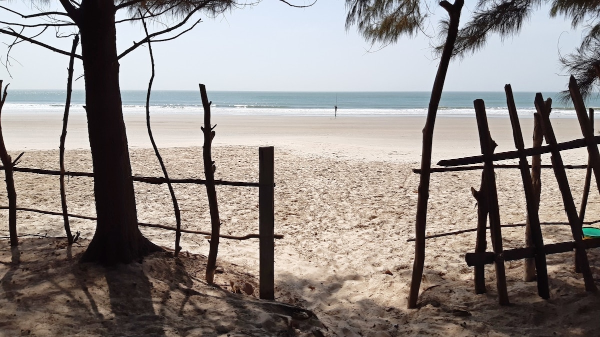 A serene view of the beach is framed by wooden fencing and trees. The sandy shore stretches towards the calm sea, where gentle waves lap at the edge. A solitary figure can be seen in the distance, adding a sense of peaceful solitude.