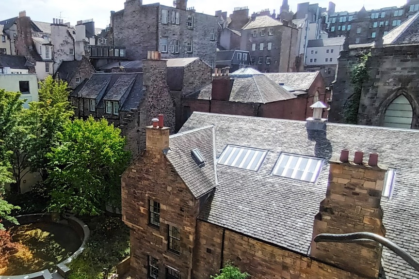 A cityscape view is presented, showcasing a collection of historic stone buildings with slate roofs. Lush greenery from nearby trees complements the architecture, while a small courtyard pond adds tranquility to the scene. The skyline reflects the charm of the Old Town.