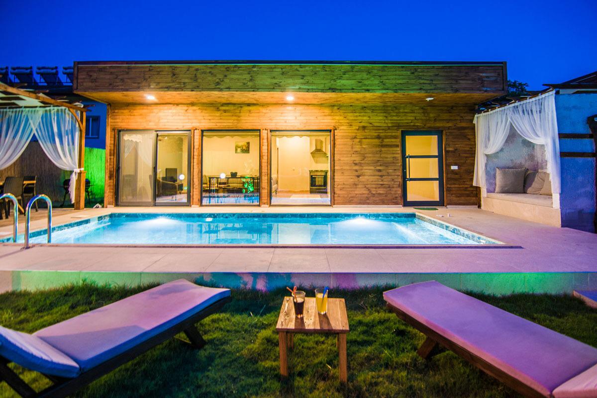 The outdoor area features a swimming pool, surrounded by lounge chairs and a small table. Soft lighting highlights the wooden structure in the background, which includes large glass doors opening to a brightly lit interior. The pool area is set within a well-manicured lawn.