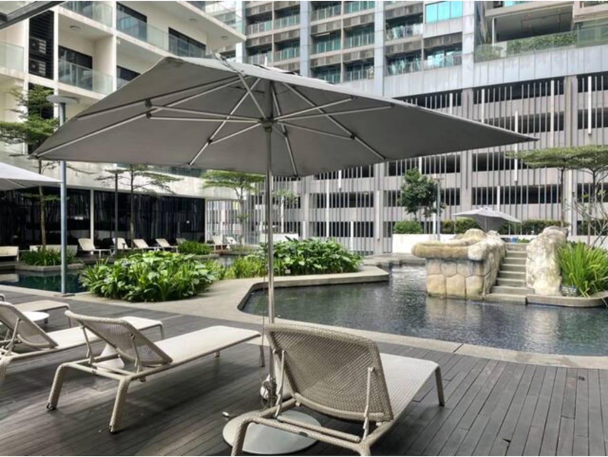 The swimming pool area is surrounded by lush greenery, featuring comfortable lounge chairs and large umbrellas for shade. The tranquil water reflects the surrounding modern architecture, while a stone feature adds an inviting touch to the space.