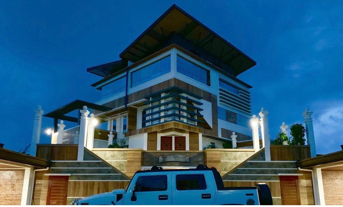 An impressive multi-story building is presented, featuring contemporary architectural elements and large glass windows. The structure is highlighted by elegant lighting, while landscaped gardens and stone steps lead to the entrance, creating a welcoming appearance against the evening sky.