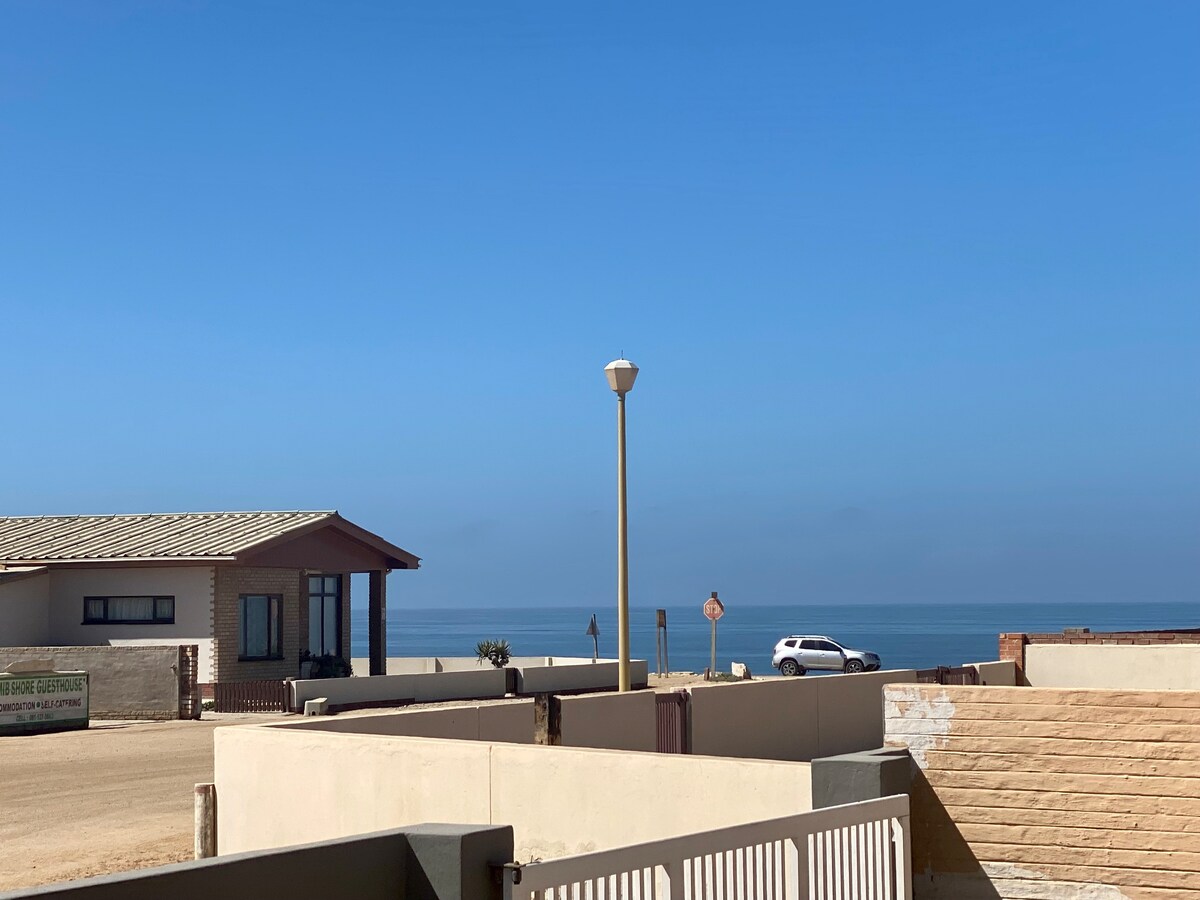 A view of the serene ocean stretches beyond the rooftops of nearby buildings, marked by clear blue skies. A residential structure with a light-colored facade is visible, along with a lamp post standing in front of the house, creating a peaceful coastal atmosphere.
