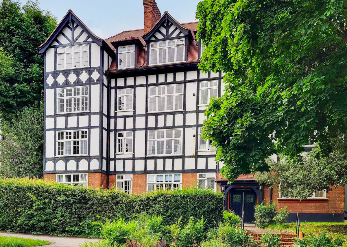 This three-story building showcases a mix of black and white timber framing, complemented by large, multi-paned windows. Lush greenery surrounds the entrance, providing a serene and inviting setting. The architectural details reflect a charming period style, enhancing the overall character of the residence.