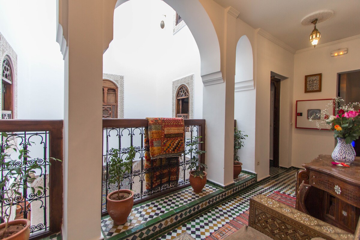 The corridor showcases intricate Moroccan tiles and features elegant arches. Potted plants line the space, with a vibrant textile draped over the railing. Natural light floods in, illuminating the traditional wooden windows visible at either end of the hallway.