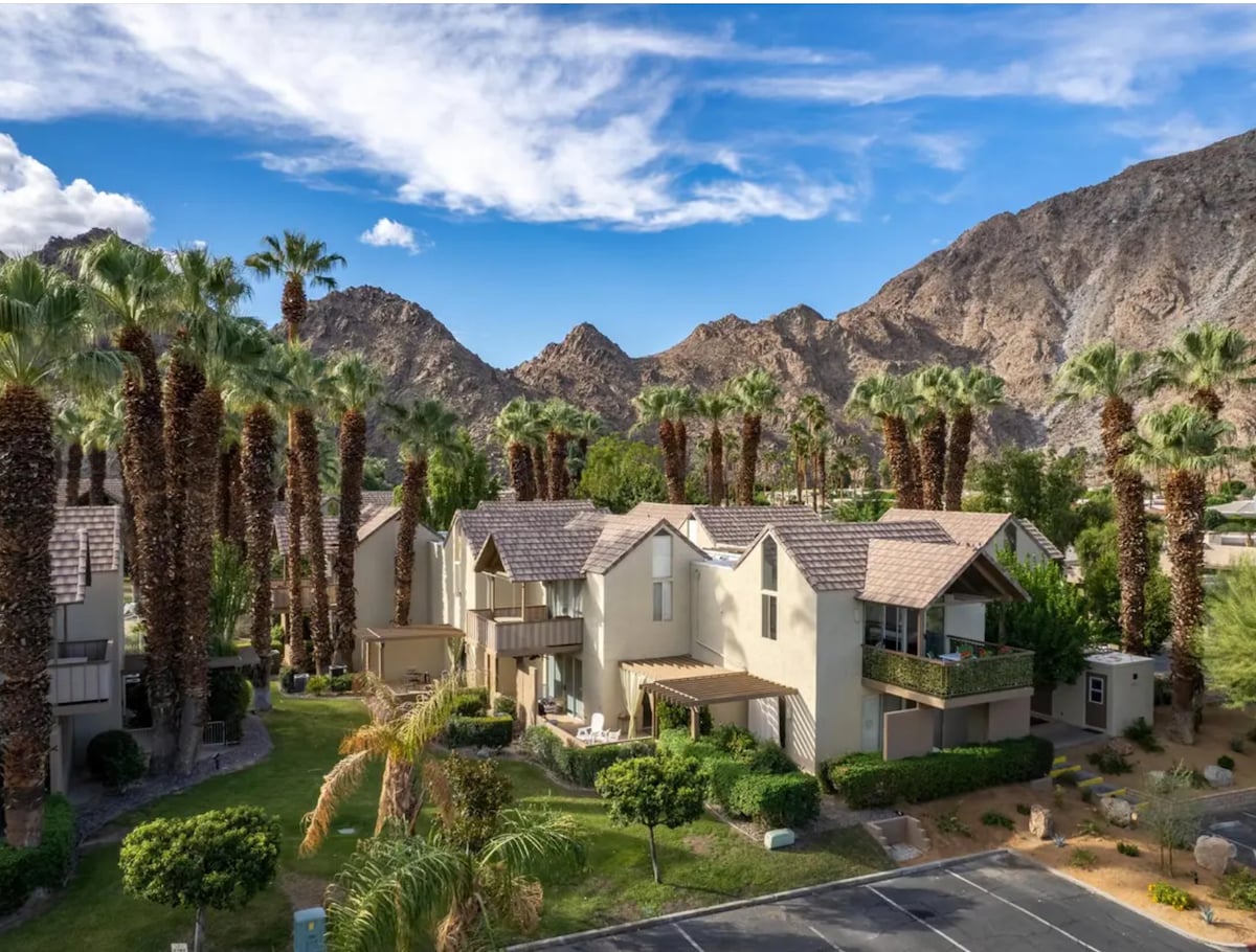 The aerial view captures a cluster of mid-century style buildings surrounded by palm trees, with stunning mountain backdrops. Lush greenery is present in the landscaping, enhancing the serene environment. The clear blue sky is dotted with scattered clouds, adding to the tranquil setting.