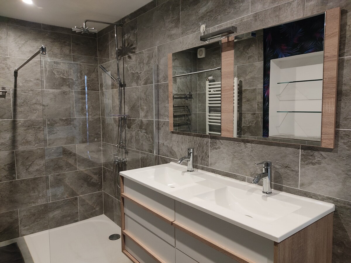 A spacious bathroom is showcased with a large double vanity featuring two sinks. A glass shower stall is positioned to one side, and a well-organized wall-mounted shelf can be seen. The walls are tiled with large gray tiles, creating a modern ambiance.
