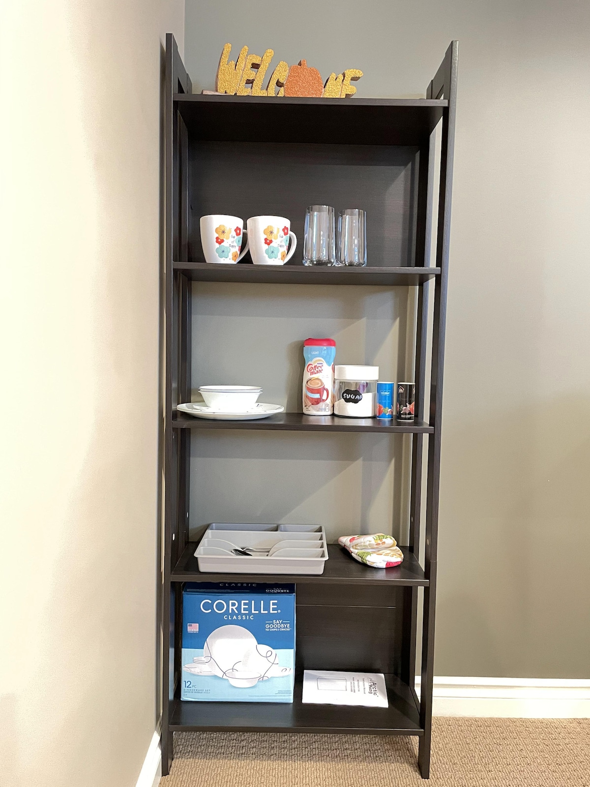 A narrow black bookshelf is displayed against a light gray wall. It holds various items: two coffee mugs, glasses, a set of bowls, a container with sugar, and a stack of cutlery. Additional items include a laundry basket and a Corelle dish set box on the bottom shelf.