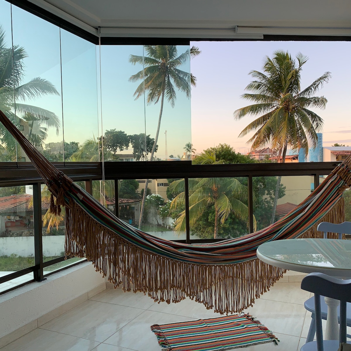 A colorful hammock hangs in a spacious balcony area, offering views of palm trees against a pastel sky. A small round table with light blue chairs is visible, alongside a striped rug on the tiled floor.