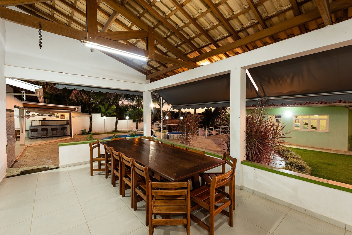 An outdoor dining area features a large wooden table surrounded by multiple chairs, set beneath a spacious roof. Outdoor lighting creates an inviting ambiance. Visible in the background are a swimming pool and landscaped greenery, enhancing the gathering space for family and friends.
