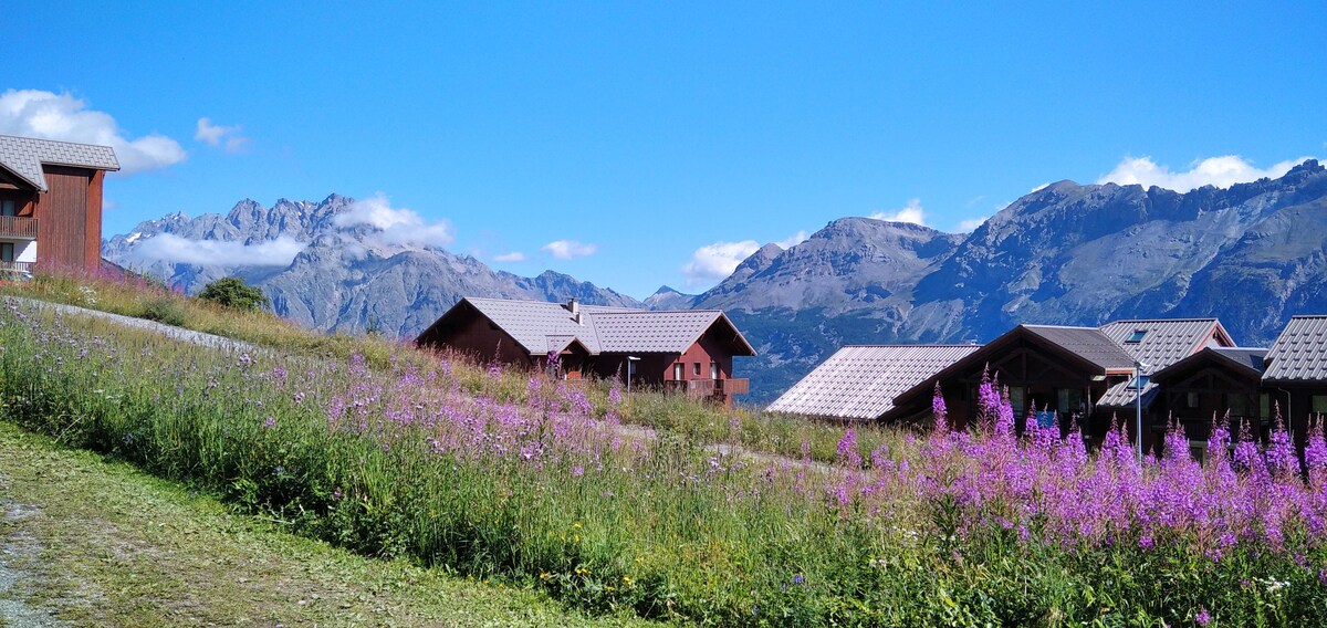 A panoramic view of the surrounding mountains is framed by colorful wildflowers in the foreground. The image features several rustic chalets with sloped roofs, harmoniously integrated into the natural landscape under a clear blue sky.