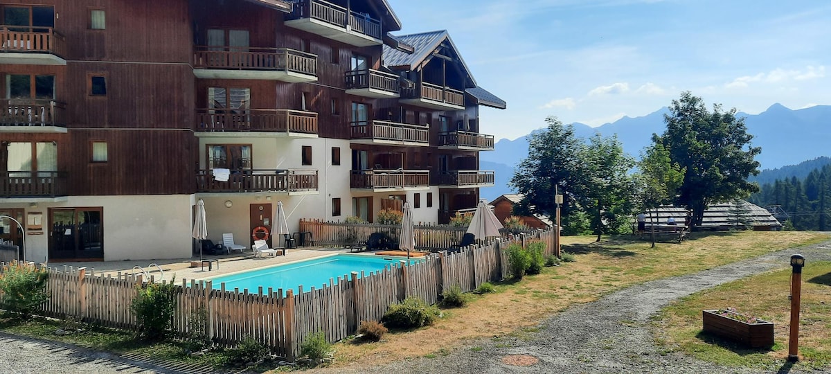 A multi-story residence is depicted with a wooden exterior and balconies. An outdoor pool is located in front, surrounded by a wooden fence. Nearby, green lawn areas and trees are visible, along with the distant mountain landscape under a clear blue sky.