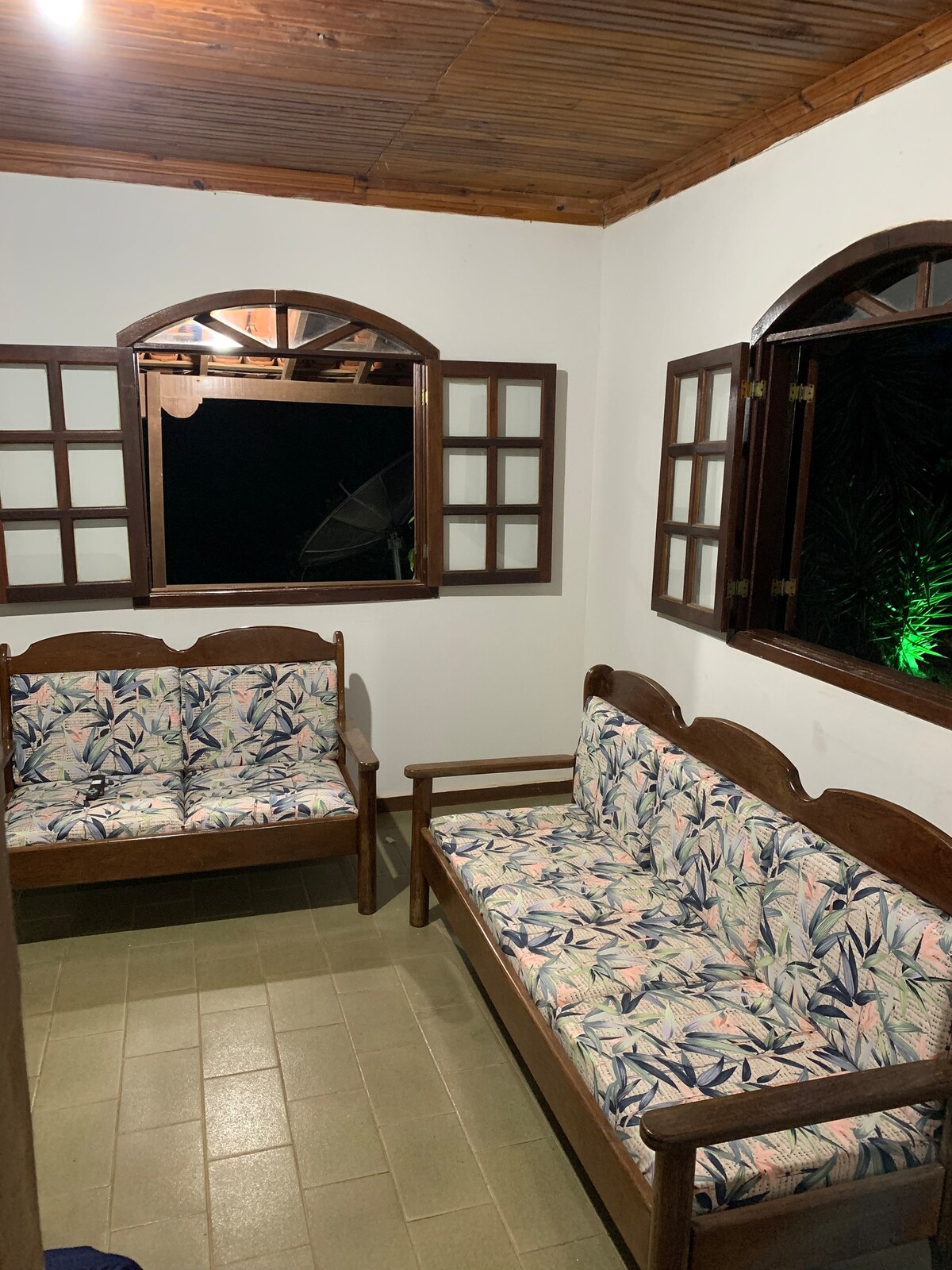 Two wooden sofas with floral-patterned cushions are arranged in a cozy sitting area, illuminated by soft lighting. Windows on either side allow for natural light, providing a glimpse of the outdoors through decorative grillwork.