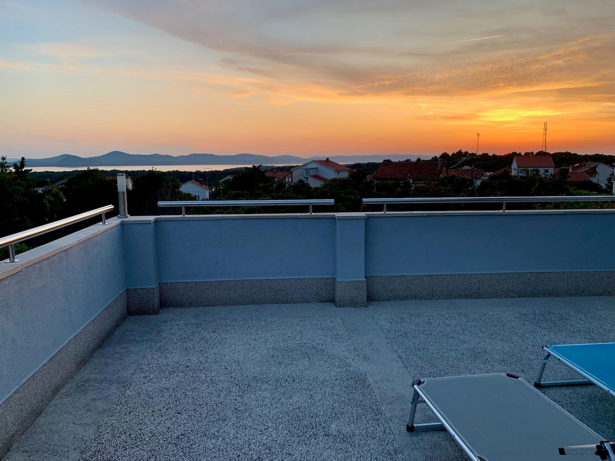 A spacious rooftop terrace is presented, featuring a smooth surface and railings. Lounge chairs are placed on one side, allowing for relaxation while enjoying the unobstructed sea views and vibrant sunset hues in the background.