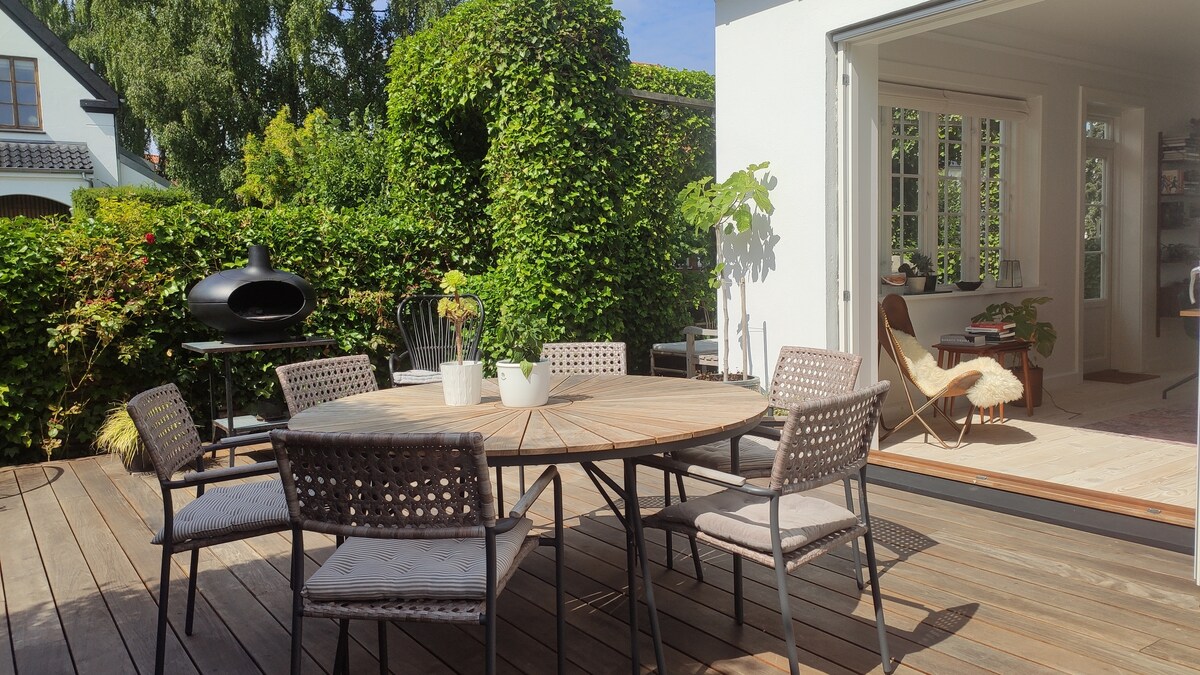 An outdoor terrace features a round wooden dining table surrounded by six woven chairs. Lush greenery provides privacy, with a grill positioned nearby. The indoor space is visible through open glass doors, showcasing a well-lit room with a workspace and natural decor.