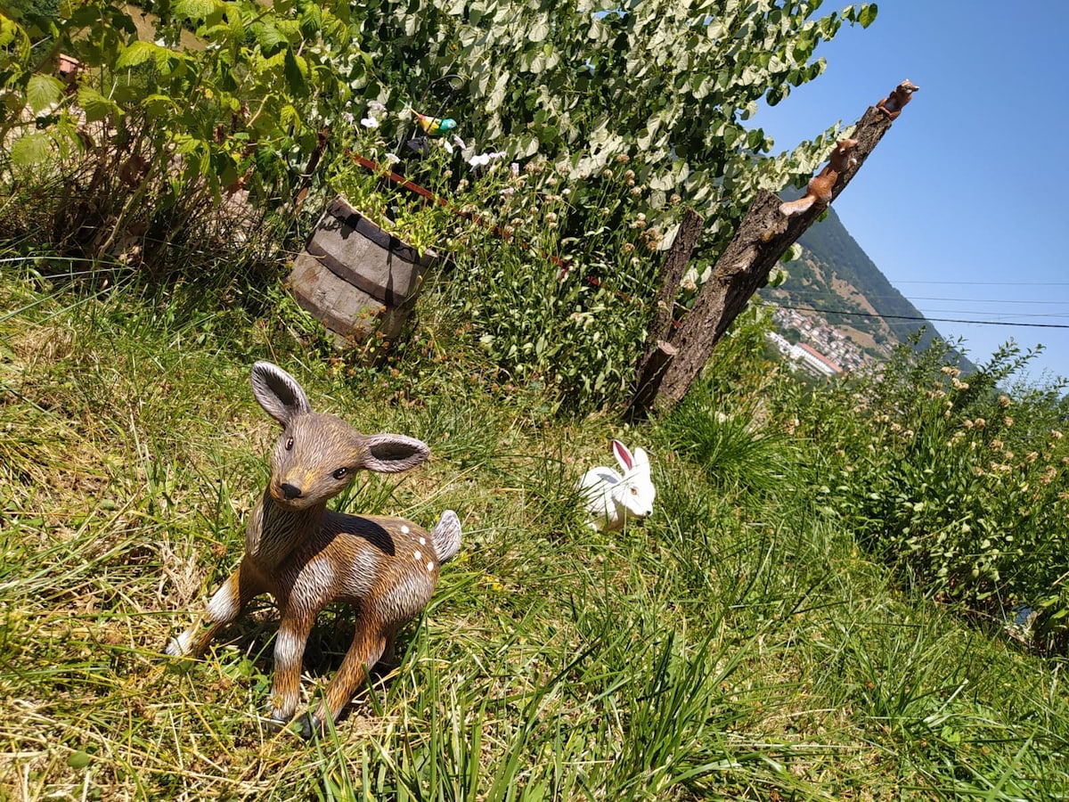 A small decorative deer figurine rests on the lush green grass, surrounded by a variety of plants. In the background, a rustic wooden barrel and a flowering plant add character to the garden, which features a scenic view of the surrounding landscape.