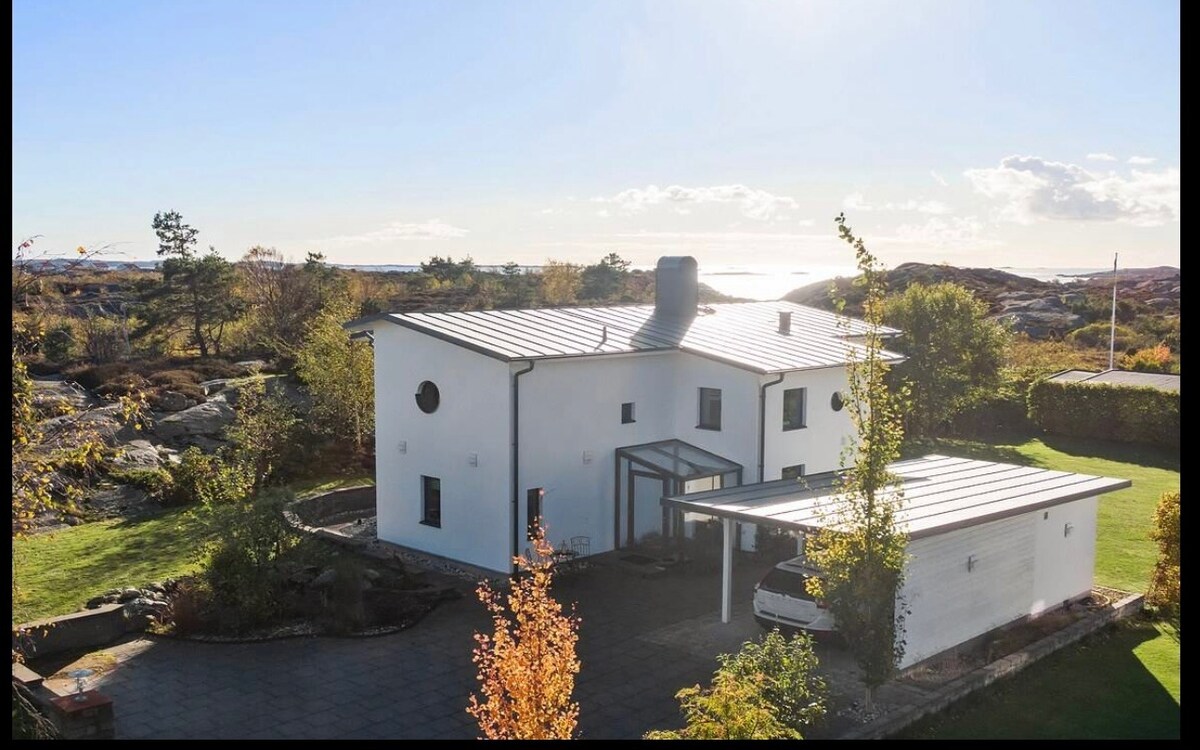 The exterior of a modern white house is displayed, featuring a silver metal roof and large windows. A circular window adds visual interest, while a covered carport is visible. Lush greenery and rocky terrain surround the property, with a view of the sea in the background.