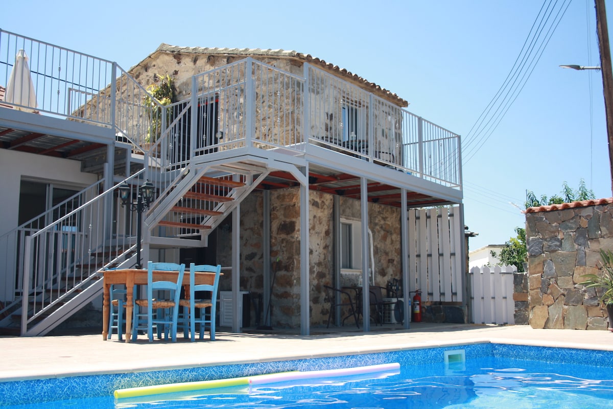 A two-story structure with a natural stone exterior is visible, surrounded by a swimming pool. A wooden dining table with blue chairs is positioned outdoors. A staircase leads to the upper floor, where a balcony is present, providing scenic views of the surroundings.
